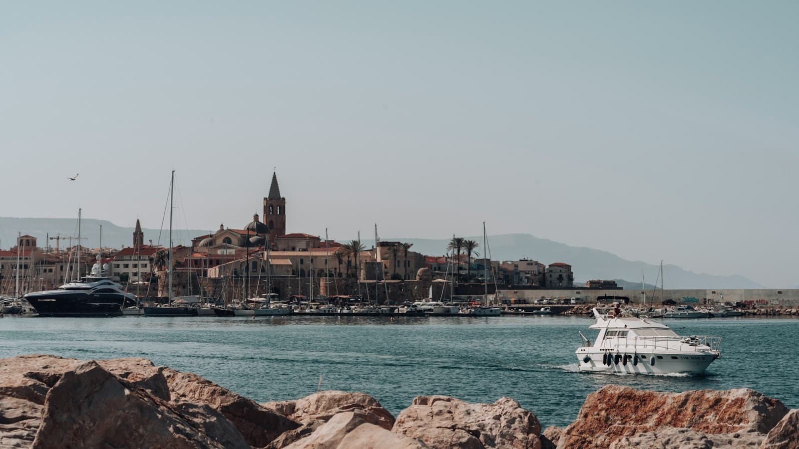 Alghero's old-town skyline across the harbour — bell tower, cathedral dome and sea walls, with boats in the foreground