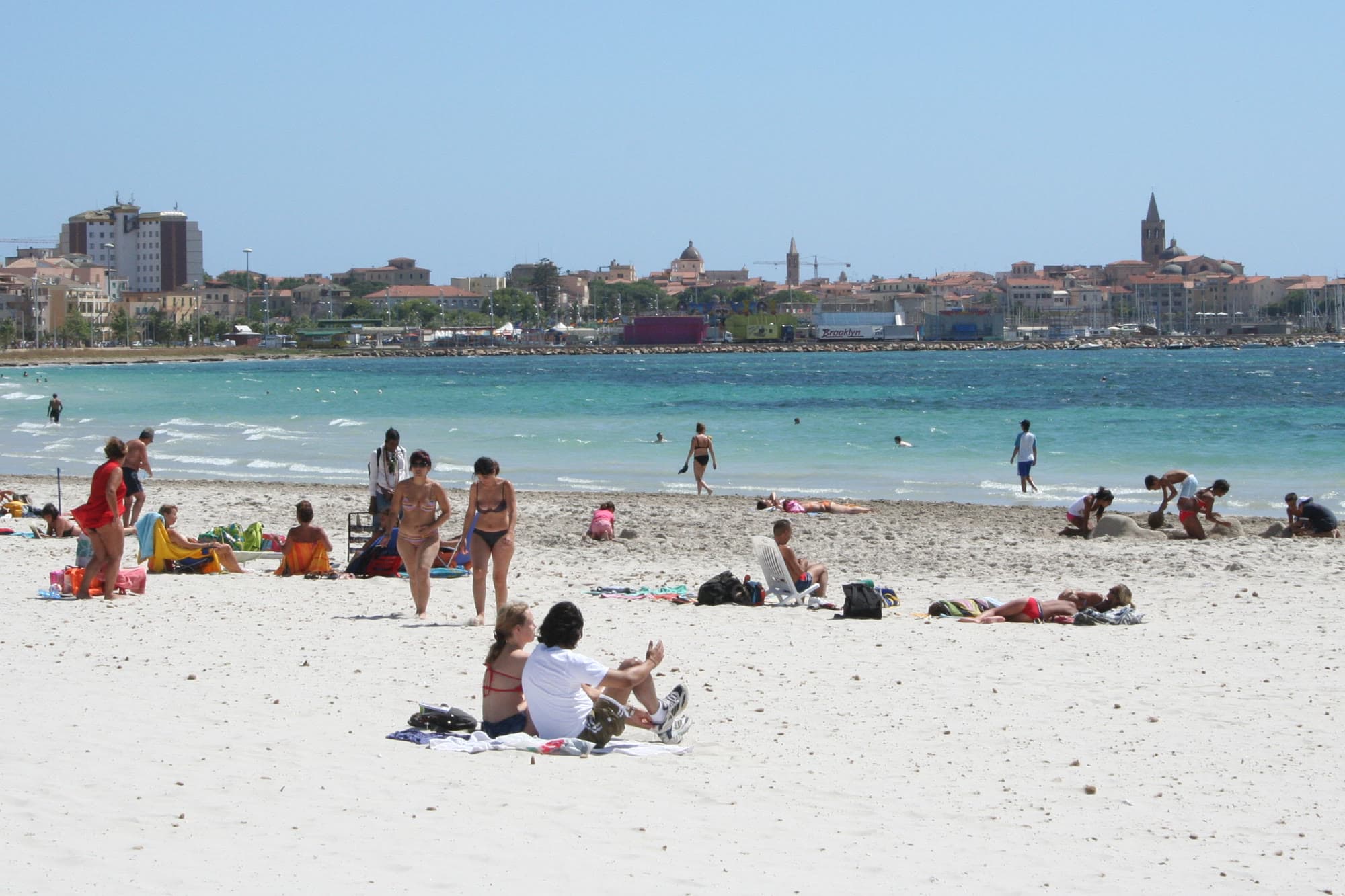 The city beach at Alghero in summer: white sand, bathers in turquoise shallow water and, across the bay, the old town's bell tower and dome